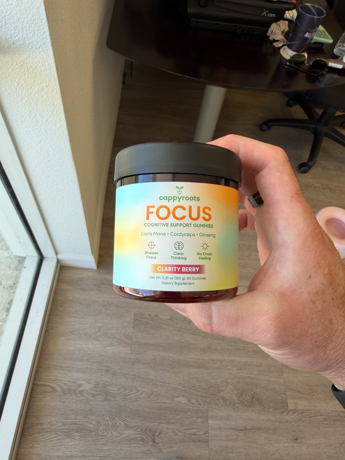 A hand holds a jar of CappyRoots Focus gummies above a wooden floor, label facing the camera